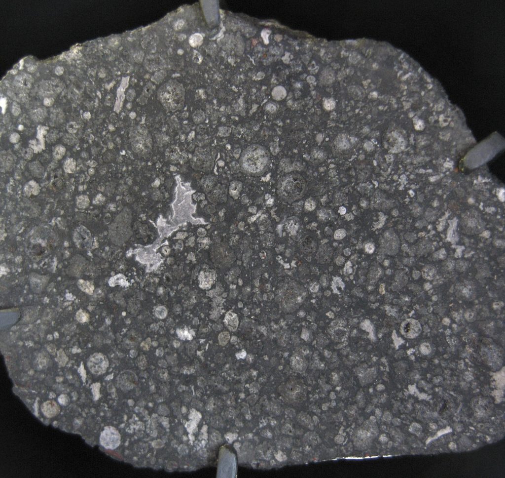Coupe polie de la météorite d'Allende : matrice gris sombre parsemée de chondres sphériques clairs et d'une inclusion blanche irrégulière.