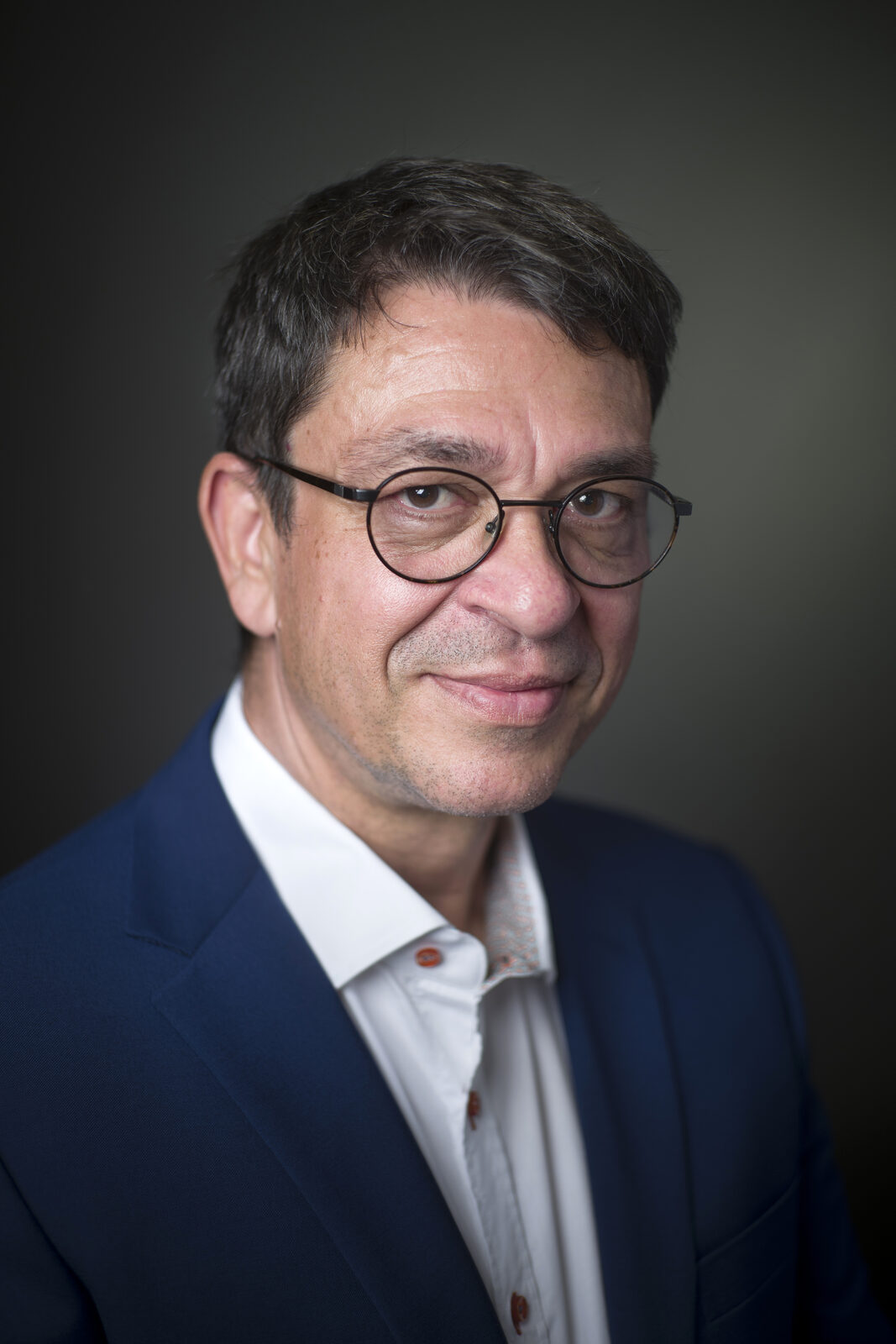 Portrait de Ramon Cortes, gemmologue, veste bleue et lunettes rondes, sur fond sombre