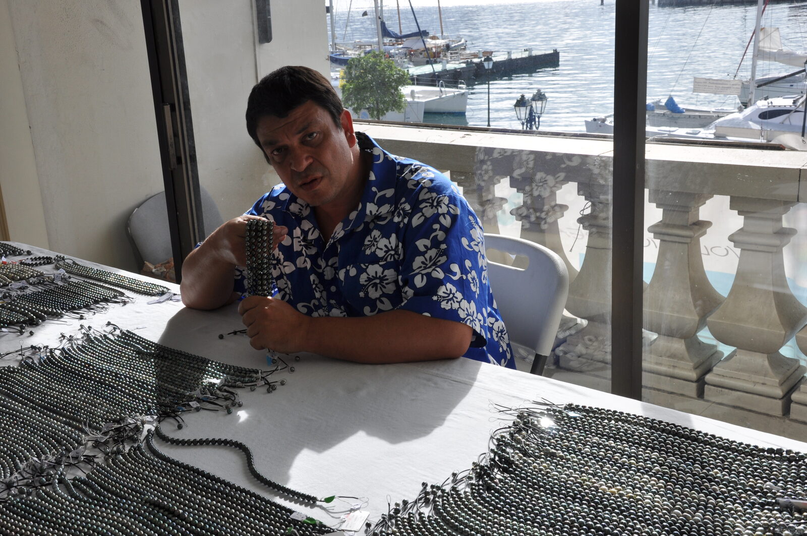 Examen d'un rang de perles de Tahiti à la centrale d'achat de Papeete, colliers étalés sur la table, port en arrière-plan