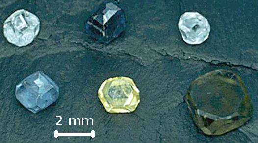 Six cristaux bruts de diamant synthétique HPHT aux formes cubo-octaédriques et aux teintes variées, avec une échelle de deux millimètres.
